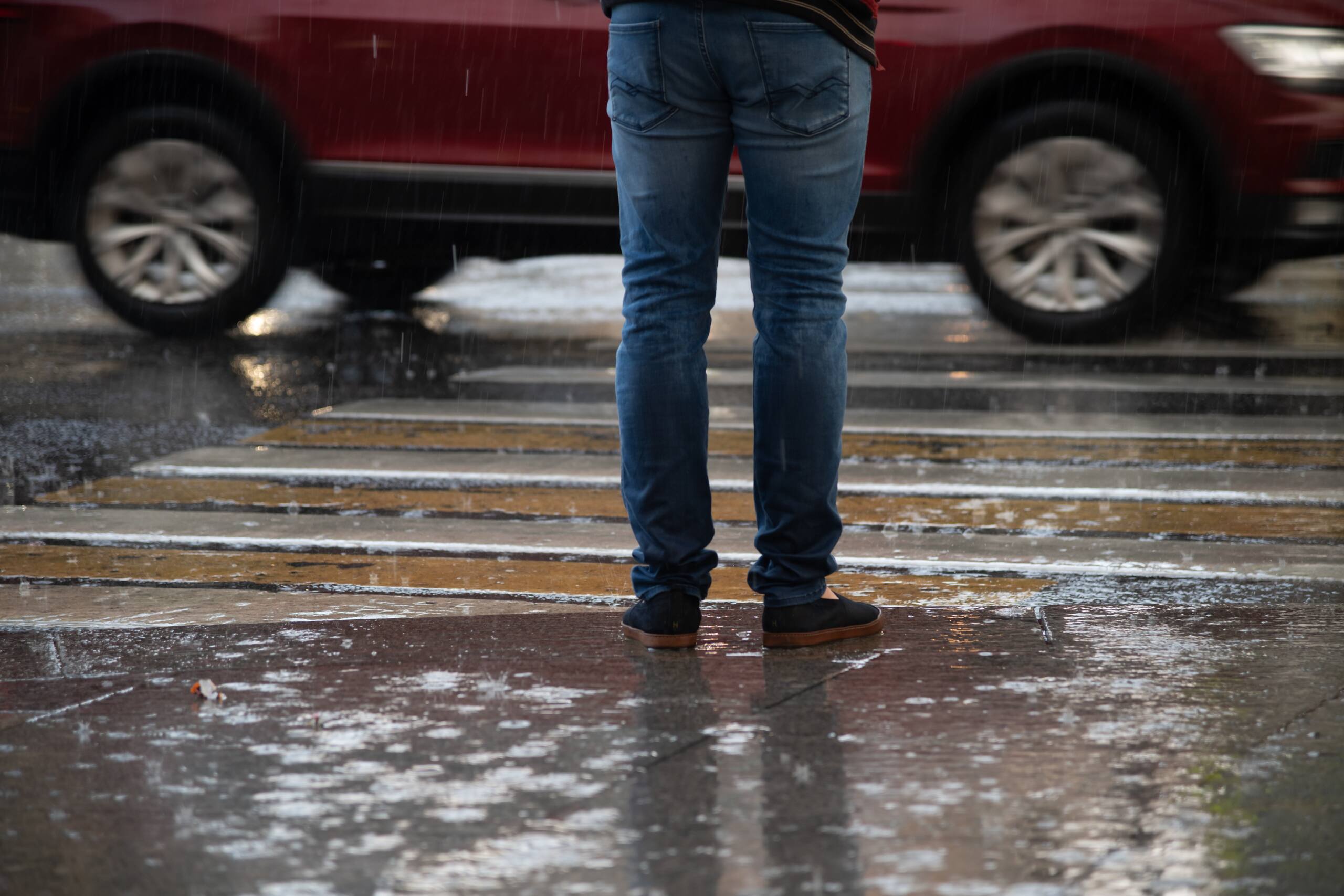 Pedestrian standing in a rainy New York crosswalk as a vehicle passes, representing no-fault insurance coverage for pedestrians without a car.