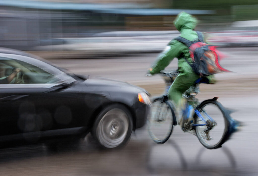 Una imagen borrosa de un coche negro chocando contra un ciclista con una mochila roja en una calle mojada.