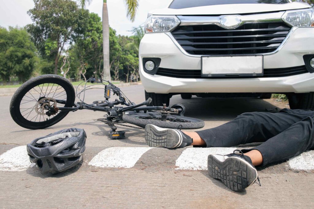 Un ciclista herido yace en el pavimento junto a su bicicleta y un casco frente a un vehículo blanco en un cruce peatonal.