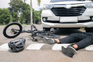 Un ciclista herido yace en el pavimento junto a su bicicleta y un casco frente a un vehículo blanco en un cruce peatonal.