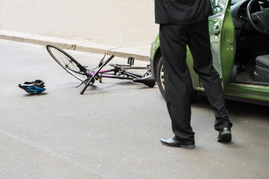 Un hombre de pie junto a la puerta abierta de un coche verde tras un accidente de dooring, con una bicicleta y un casco en el suelo.