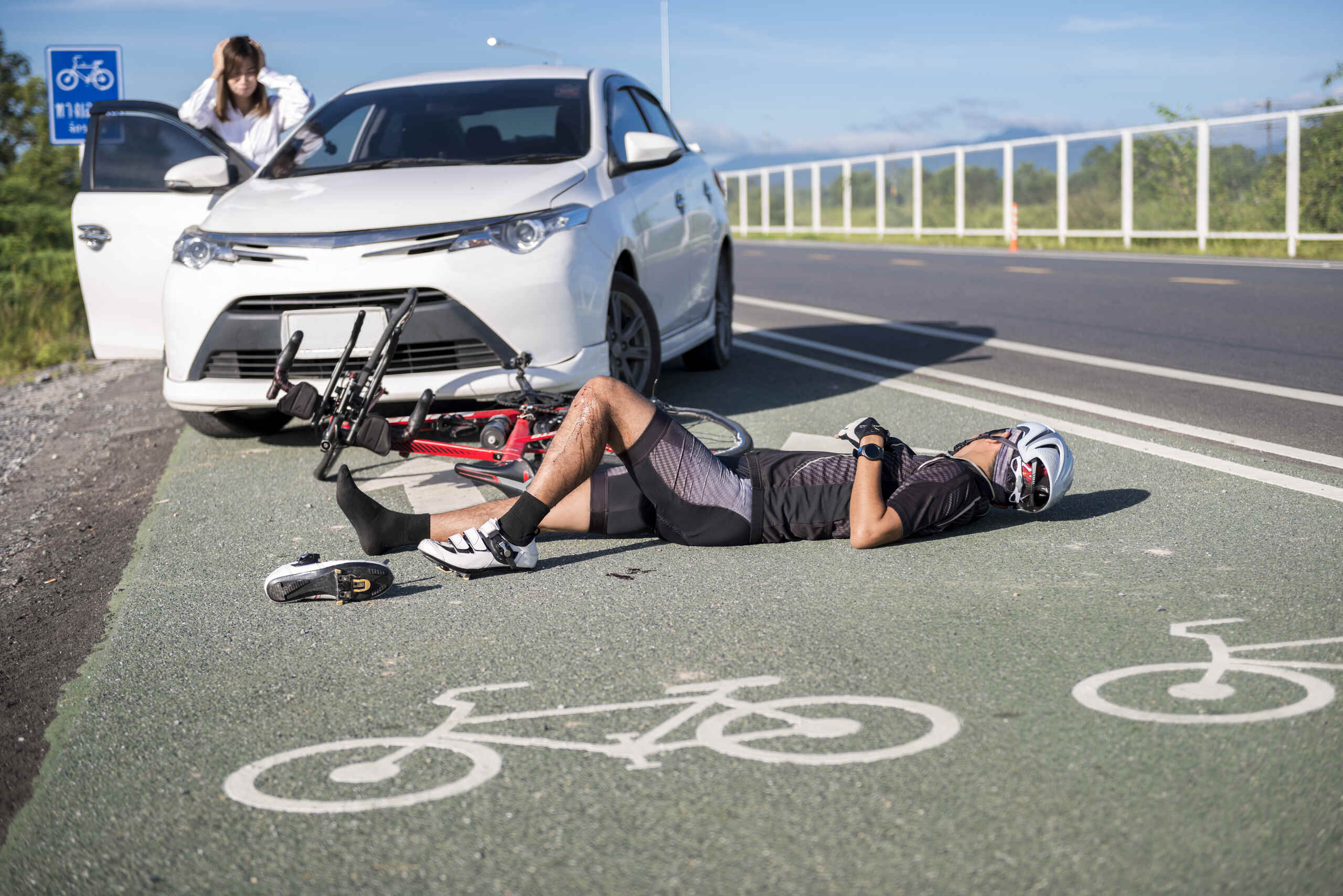 Un ciclista lesionado tendido en un carril bici verde, con su bicicleta destrozada delante de un coche blanco tras una colisión.