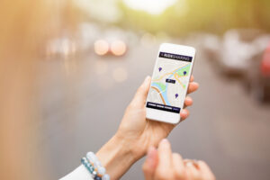 A person sits at a desk with an open laptop, holding a smartphone that displays the Uber and Lyft mobile app icons.