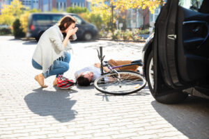 Una mujer angustiada se arrodilla junto a un ciclista herido en el suelo tras un accidente con un coche negro en un estacionamiento.