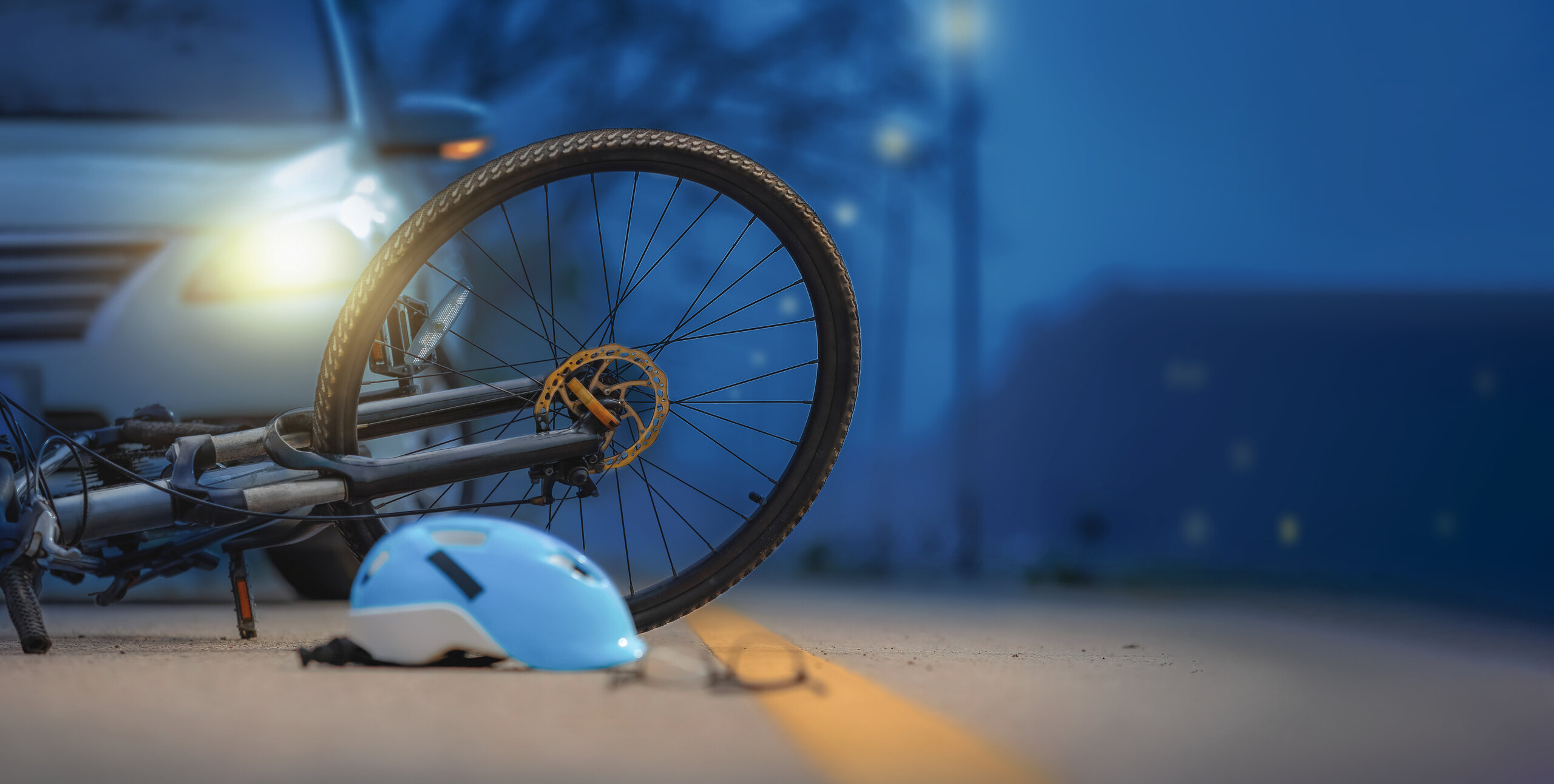 Una bicicleta y un casco azul yacen en el pavimento por la noche frente a los faros encendidos de un coche tras un accidente.