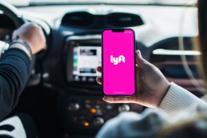 A person inside a vehicle holds a smartphone displaying the Lyft mobile app icon on a bright pink screen.