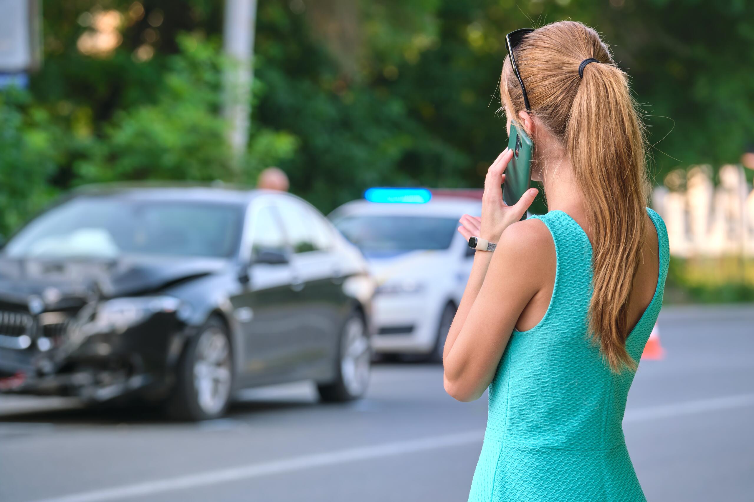 A woman in a teal dress stands by the roadside making a phone call next to a black car with significant front-end damage.