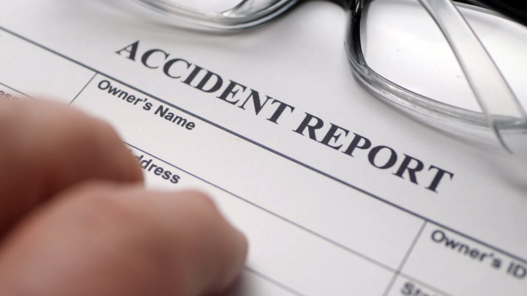 Hand filling out an official accident report form next to reading glasses demonstrating NY construction accident settlements realistic ranges by injury type.