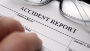 Hand filling out an official accident report form next to reading glasses demonstrating NY construction accident settlements realistic ranges by injury type.